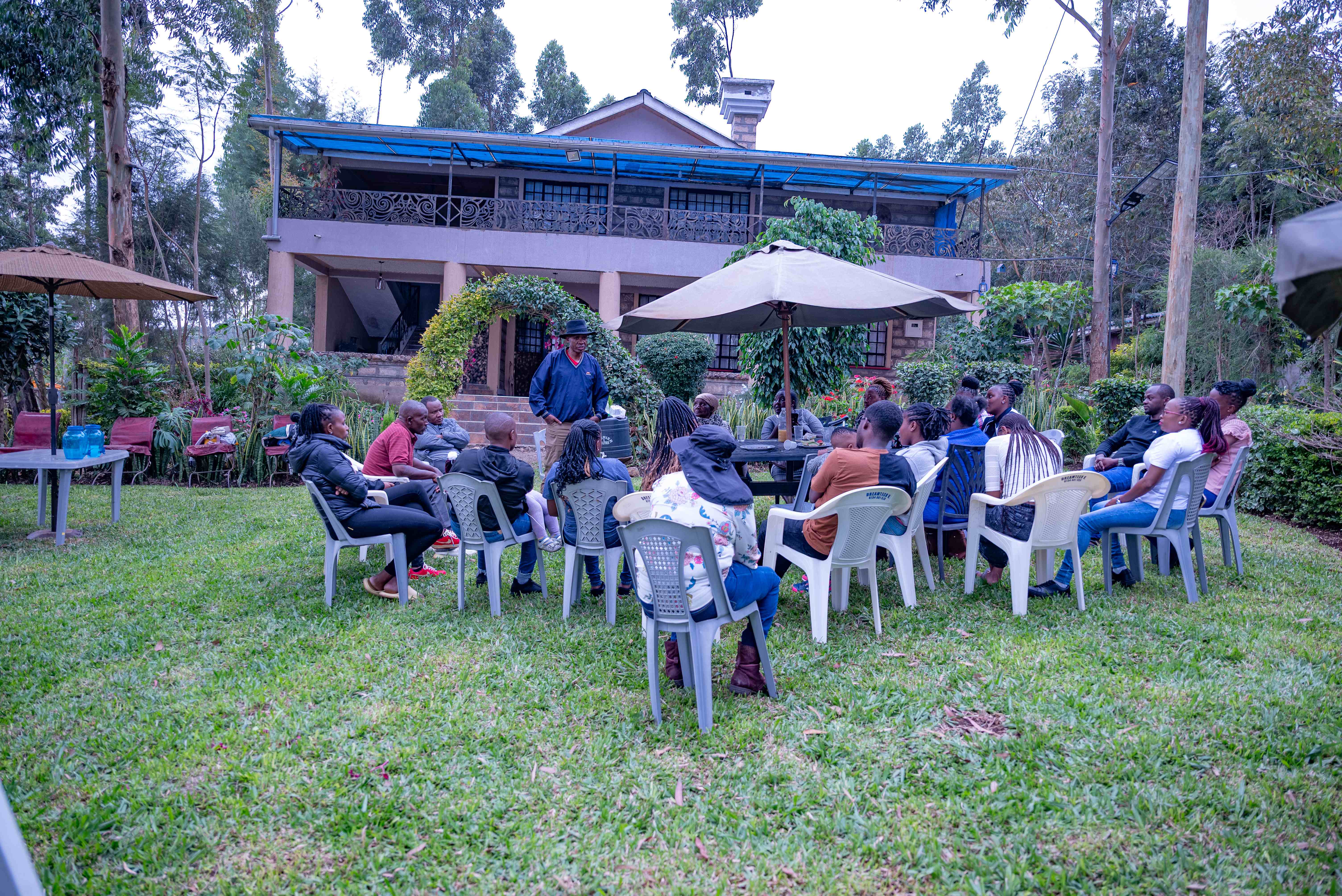 Large family gathering with people seated in a semi-circle on the farm lawn, with a facilitator leading the discussion in front of the beautiful main farmhouse